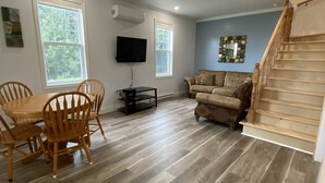 A spacious living room with a wooden dining table and chairs, a sofa, a TV mounted on the wall, and a staircase leading to another level. - New! Fabulous Farmhouse (Cape Dauphin)