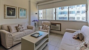 En stue med en beige sofa, en hvid lænestol, et sofabord og et vindue med udsigt til bygninger.