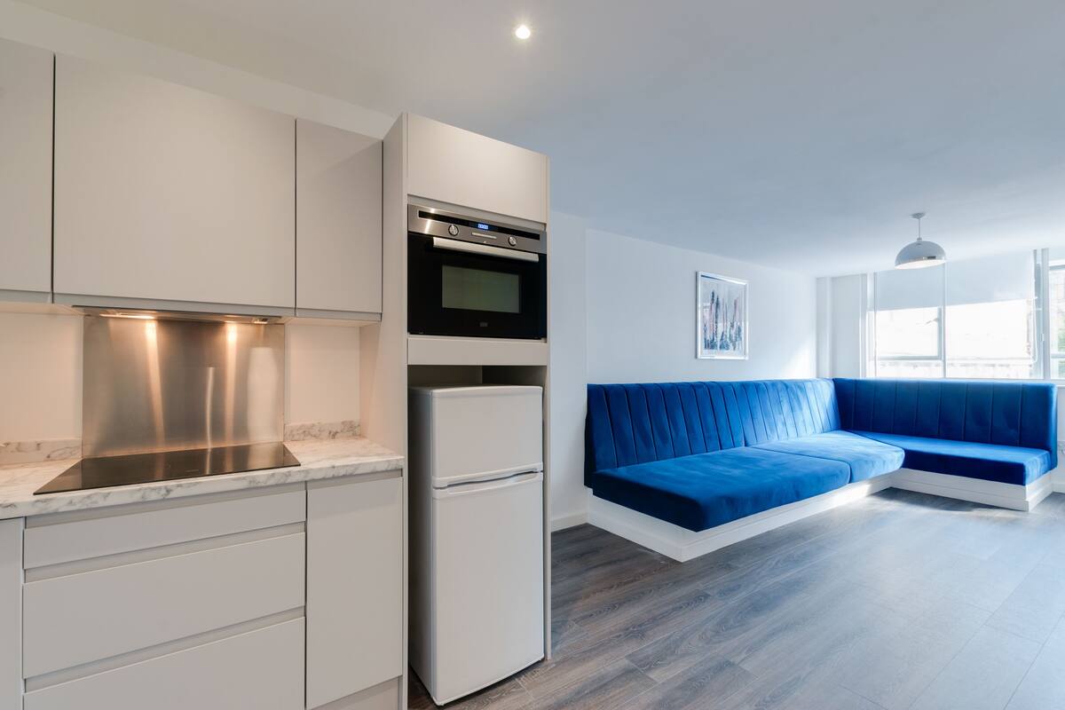 A modern kitchen with white cabinetry, a built-in oven, and a refrigerator. There is a blue sofa in the living area with a window providing natural light.