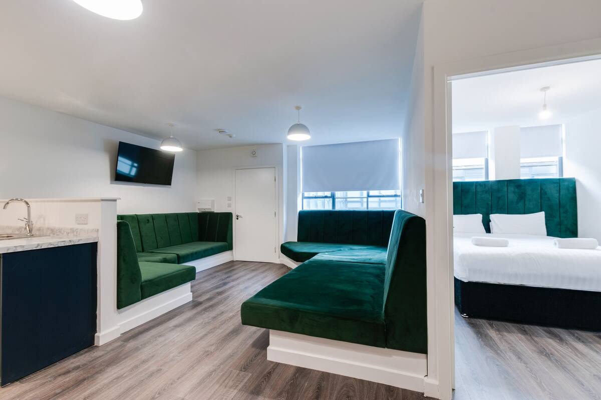 A modern hotel room with a green sofa, a bed with white bedding, a flat-screen TV mounted on the wall, and a wooden floor.