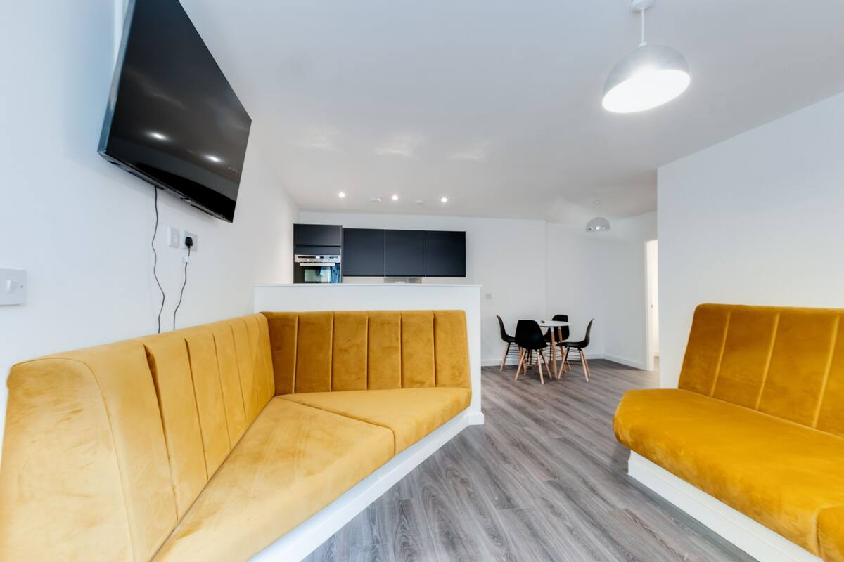 A modern living room with a sofa, a flat-screen TV mounted on the wall, a dining area with a table and chairs, and a kitchen area with cabinets.