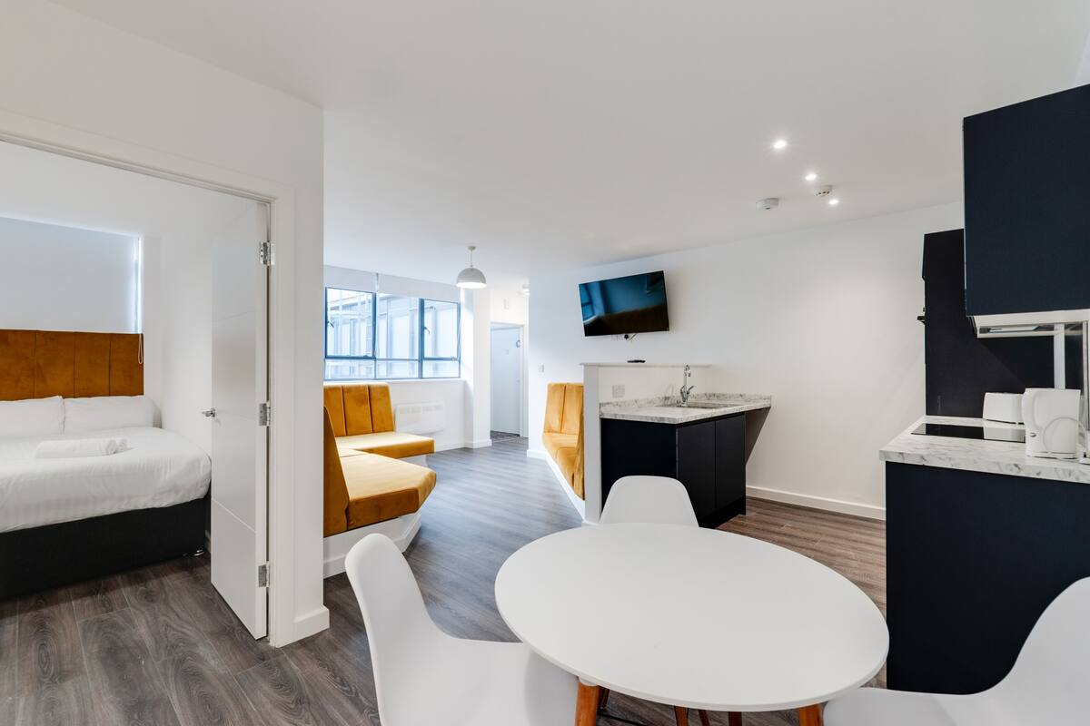 A modern hotel room with a bed, a dining area with a table and chairs, a kitchen area, and a large window providing natural light.