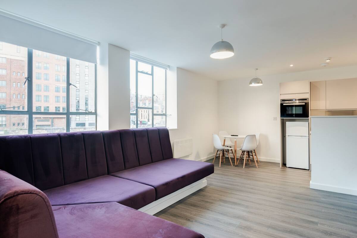 A modern apartment with a large window, a sofa, a dining area, and a kitchen.