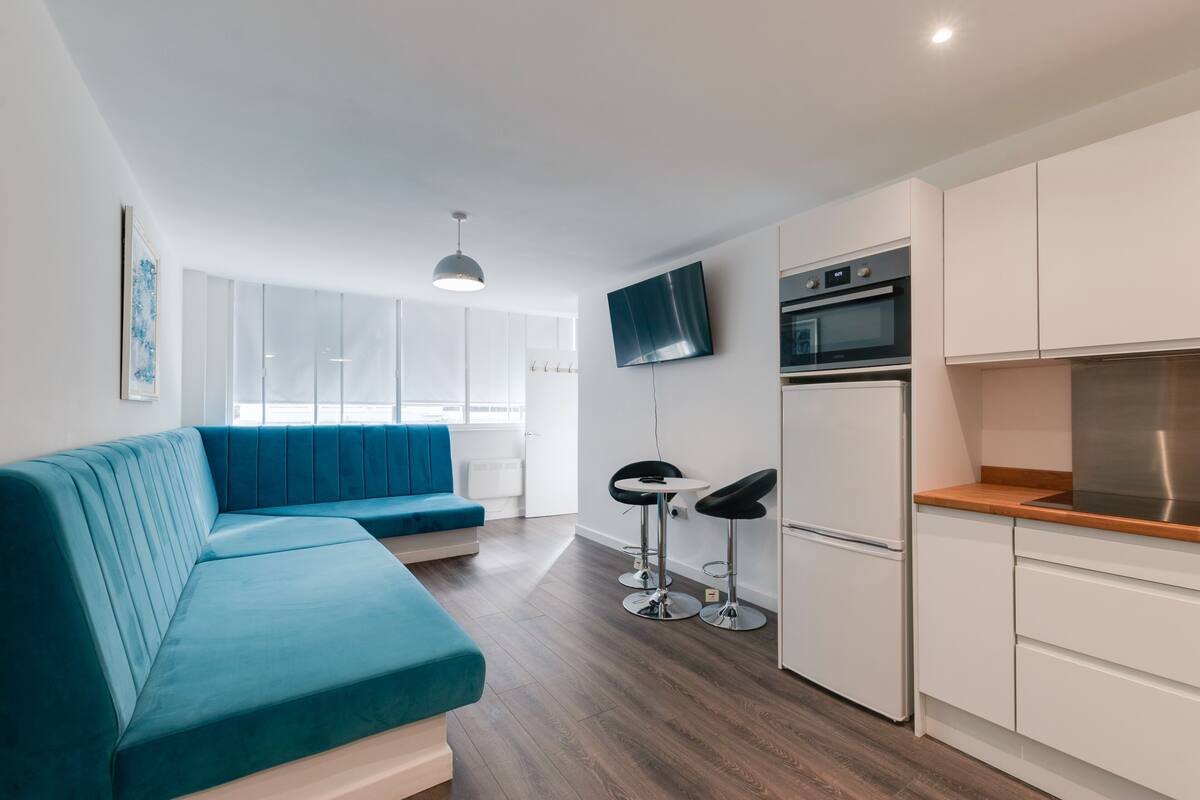 A modern kitchen with white cabinets, a built-in oven, and a stainless steel range hood. There is a blue sofa and two black bar stools in the living area.