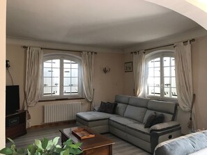 Een woonkamer met een grijze bank, een houten salontafel en een tv-meubel. Er zijn twee ramen met gordijnen, een radiator en een wandlamp.