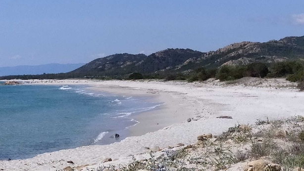 A beach with clear blue water, sandy shore, and rocky hills in the background.