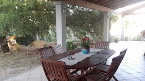An outdoor patio with a wooden table and chairs, surrounded by potted plants and a tree.