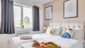 A hotel room with a bed, a window with curtains, a bedside table, and a wall with two framed pictures. - City Centre Flat - Free Parking, Wifi & SmartTV (Milton Keynes)