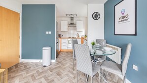 A modern kitchen with white cabinets, a glass dining table, and a blue accent wall. - City Centre Flat - Free Parking, Wifi & SmartTV (Milton Keynes)