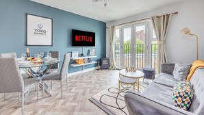A modern living room with a glass dining table, grey upholstered chairs, a grey sofa, and a television displaying 'NETFLIX'. - City Centre Flat - Free Parking, Wifi & SmartTV (Milton Keynes)