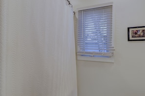 A bathroom with a shower curtain, a window with blinds, and a framed picture on the wall.