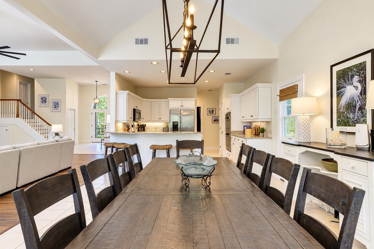 A spacious dining area with a long wooden table and chairs, a kitchen with white cabinets, and a staircase leading to an upper level.