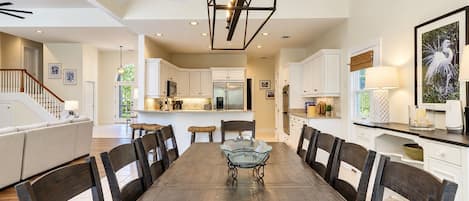 A spacious dining area with a long wooden table and chairs, a kitchen with white cabinets, and a staircase leading to an upper level.