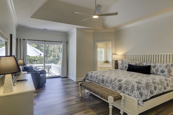 A bedroom with a bed, a ceiling fan, a nightstand, a lamp, a bench, and a view of the outdoors through a sliding glass door.