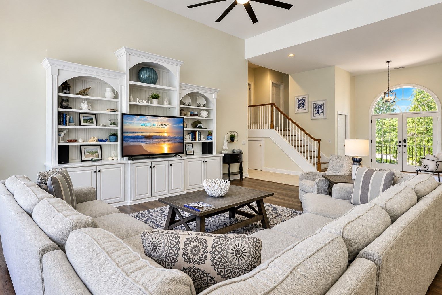 A spacious living room with a large flat-screen TV, a fireplace, a staircase, and a view of greenery through a large window.