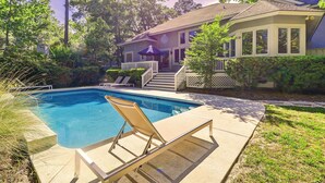 A residential swimming pool with a lounge chair, surrounded by a well-maintained lawn and a house with a porch.