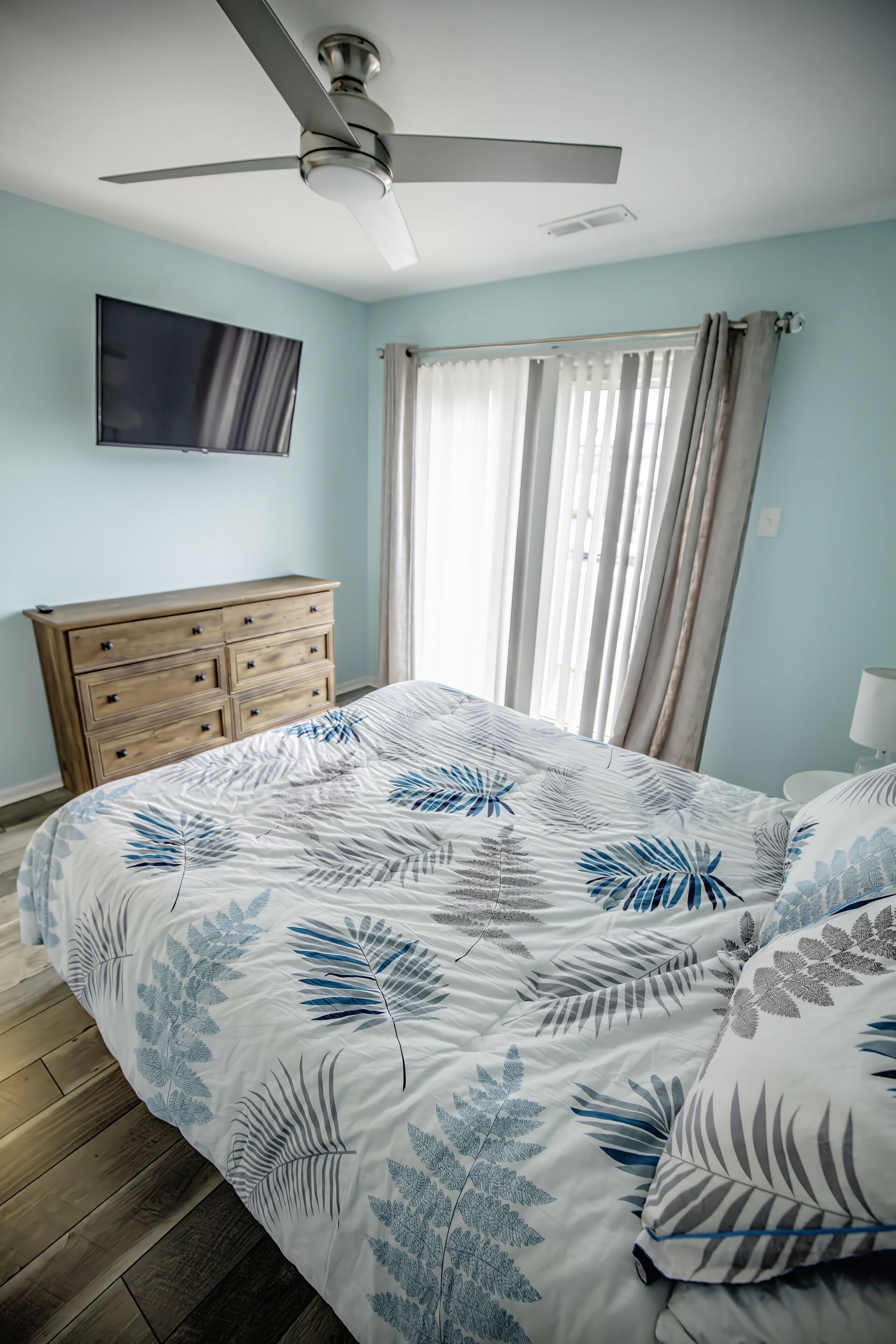 A bedroom with a bed, a wooden dresser, a ceiling fan, a TV mounted on the wall, and a window with curtains.