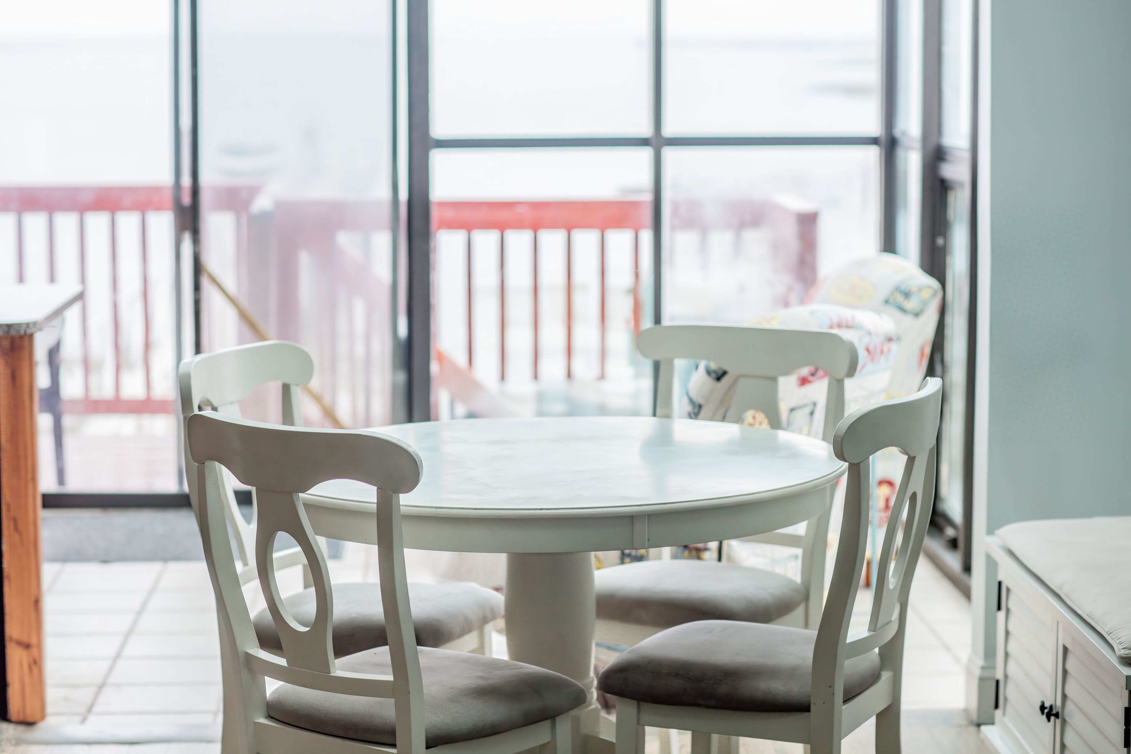 A round table with white chairs and a glass top, positioned in a room with large windows.