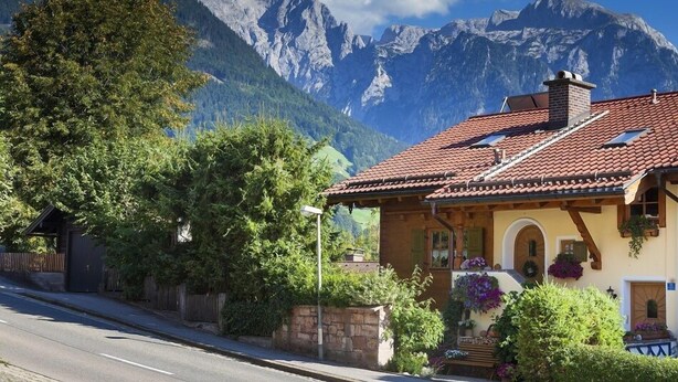 Un chalet traditionnel avec un toit recouvert de tuiles rouges, des poutres en bois et un mur en pierre. Il possède un petit porche agrémenté de plantes en pot et offre une vue imprenable sur les montagnes.