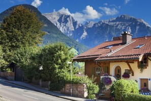 A traditional chalet with a red-tiled roof, wooden beams, and a stone wall. It features a small porch with potted plants and a mountain backdrop. - Ferienwohnung Watzmann, für 1-4 Personen, 54 qm, Südterrasse, Garage, Separater Eingang (Berchtesgaden)