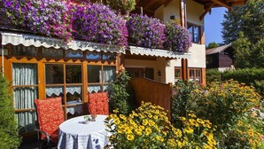 Een traditionele chalet met een balkon vol paarse bloemen, een houten terras en een tafel met stoelen.