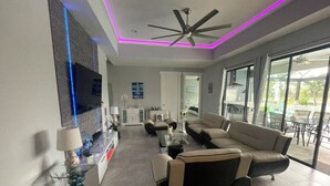 A modern living room with a sofa, TV, and ceiling fan. - South-facing Waterfront Villa 3BR w/ Heated Pool & Spa (Cape Coral)