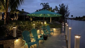 A waterfront patio with chairs and a table under a green umbrella, surrounded by a brick wall and palm trees. - South-facing Waterfront Villa 3BR w/ Heated Pool & Spa (Cape Coral)