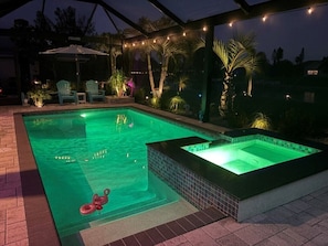 A swimming pool with a hot tub, surrounded by chairs and string lights.