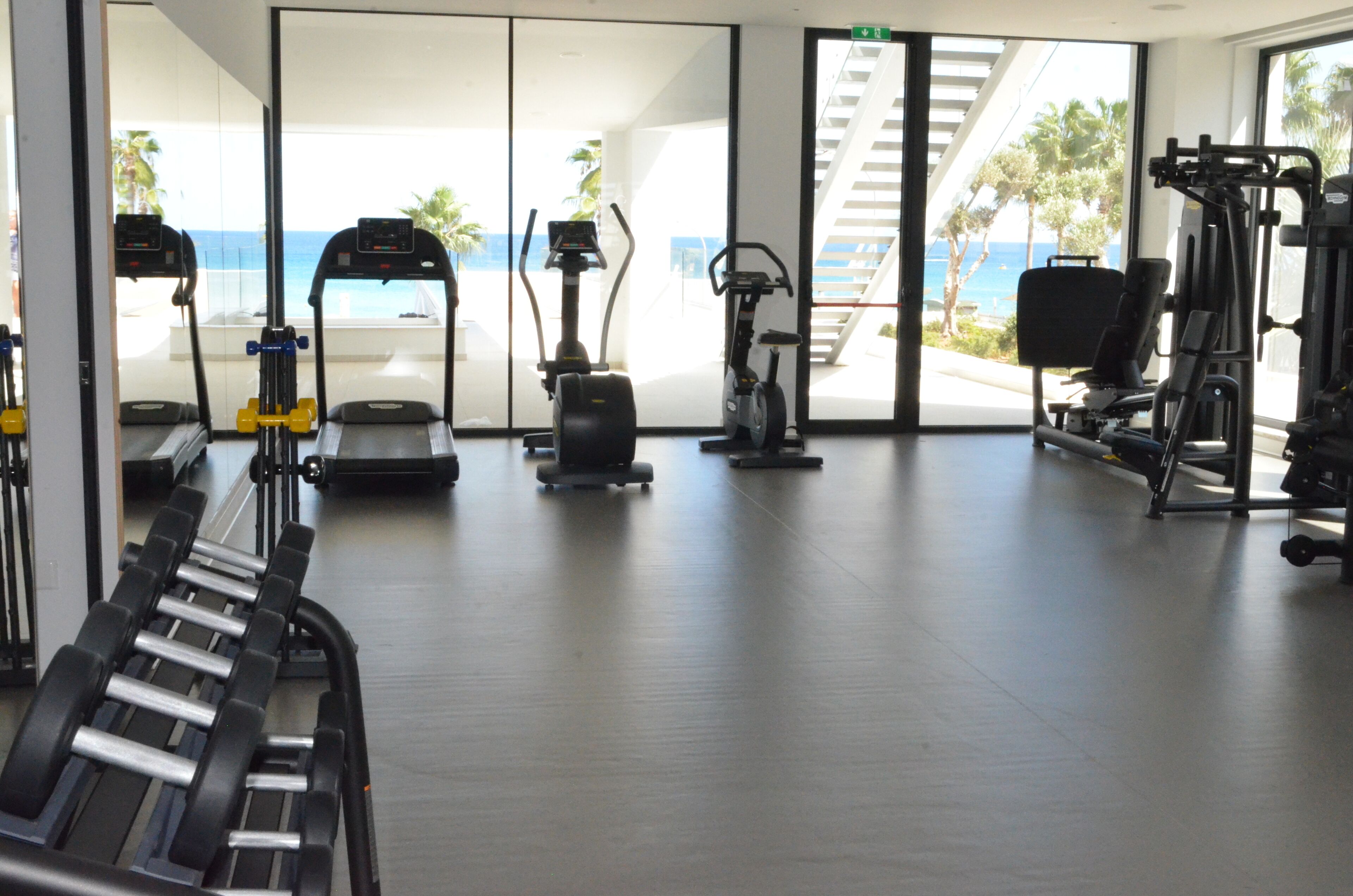A gym with cardio machines, weightlifting equipment, and a view of the ocean through large windows.