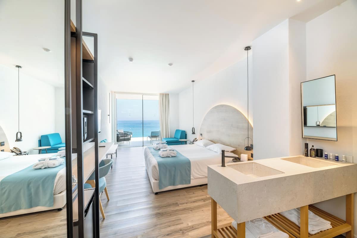 A modern hotel room with two beds, a large bathroom with a sink, and a view of the sea.