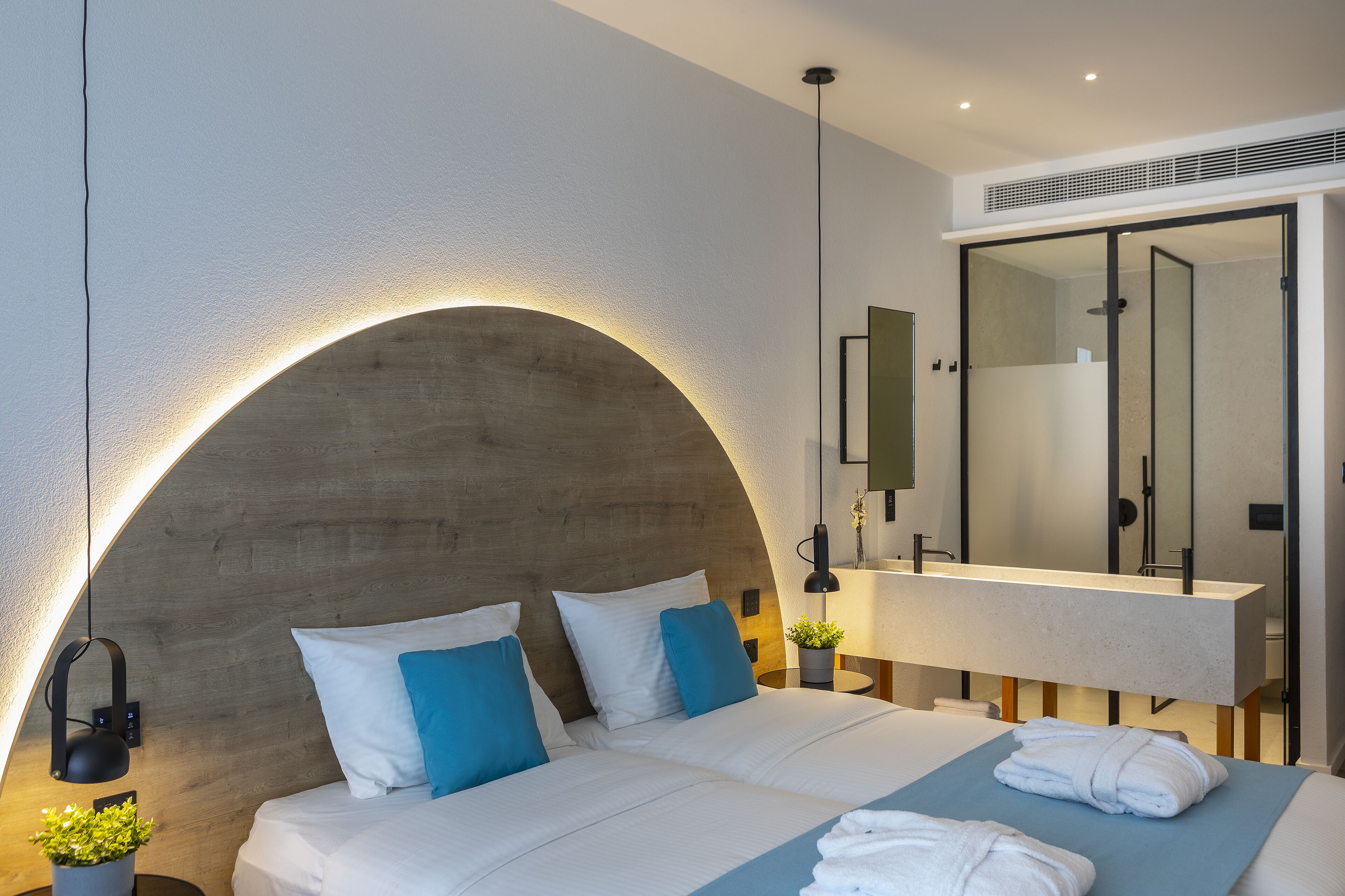 A modern hotel room with a curved headboard, a large mirror, and a neatly made bed with white linens and blue pillows.