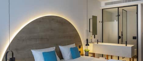 A modern hotel room with a curved headboard, a large mirror, and a neatly made bed with white linens and blue pillows.