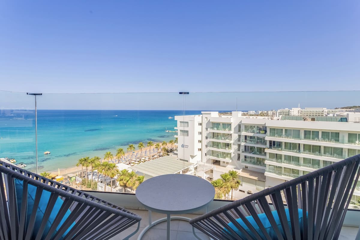 A balcony with a view of the sea, a pool area, and modern buildings.