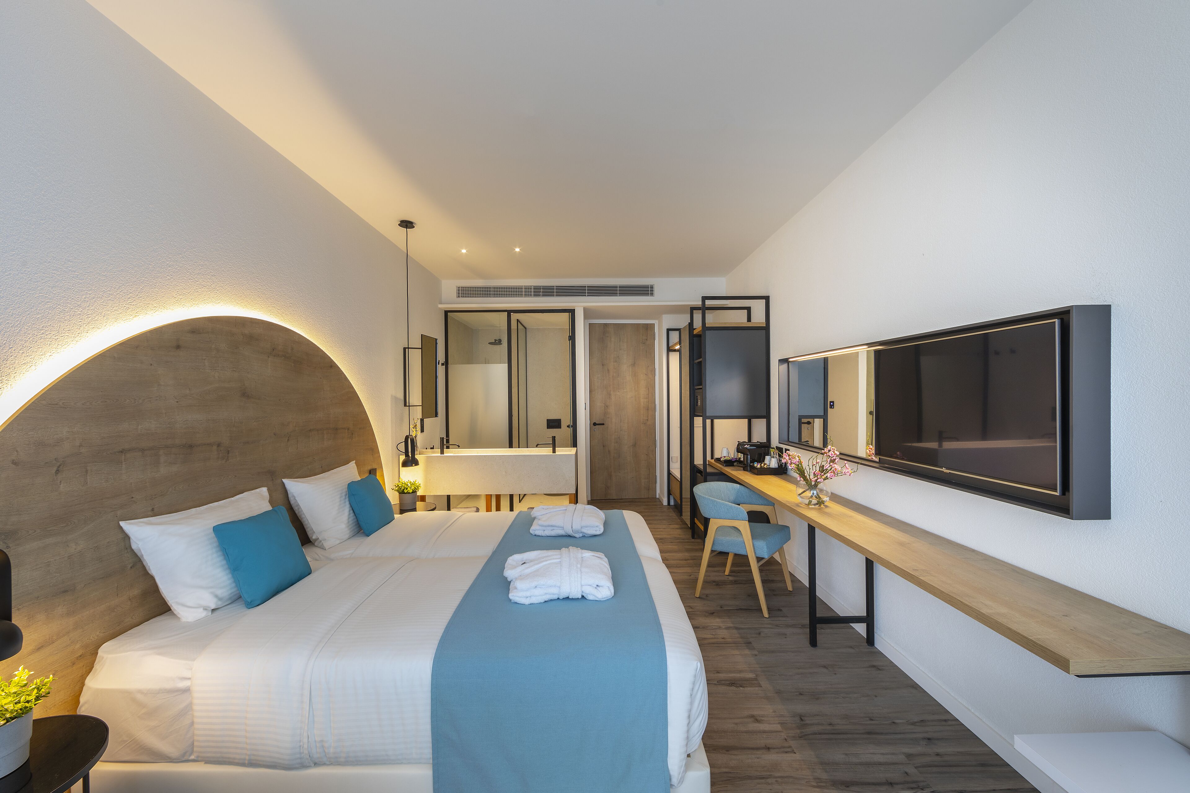 A modern hotel room with a large bed, a desk with a chair, a TV, and a bathroom visible through an open door.