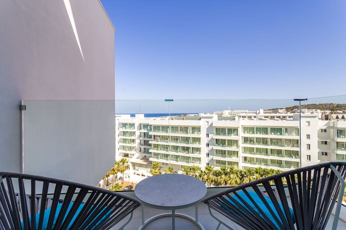 A balcony with two chairs and a small table, overlooking a modern building complex and the sea.