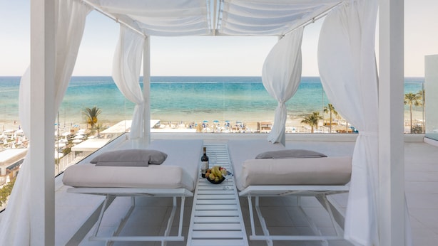 A balcony with two white cushioned chairs, a table with a bottle and fruit, and a view of the beach and ocean. - Flamingo Paradise Beach Hotel Adults Only (Paralimni)