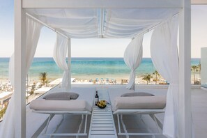 A balcony with two white cushioned chairs, a table with a bottle and fruit, and a view of the beach and ocean. - Flamingo Paradise Beach Hotel Adults Only (Paralimni)