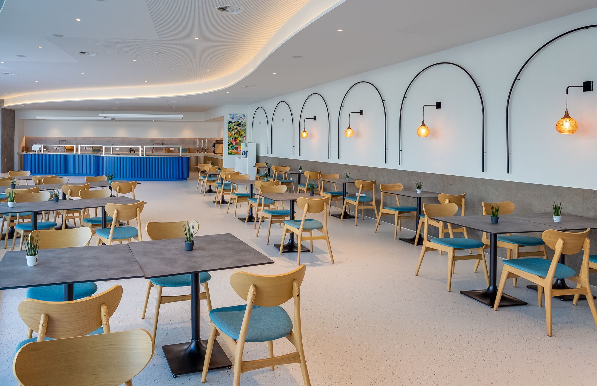 A spacious restaurant with multiple tables and chairs, a kitchen area in the background, and wall-mounted lighting fixtures.