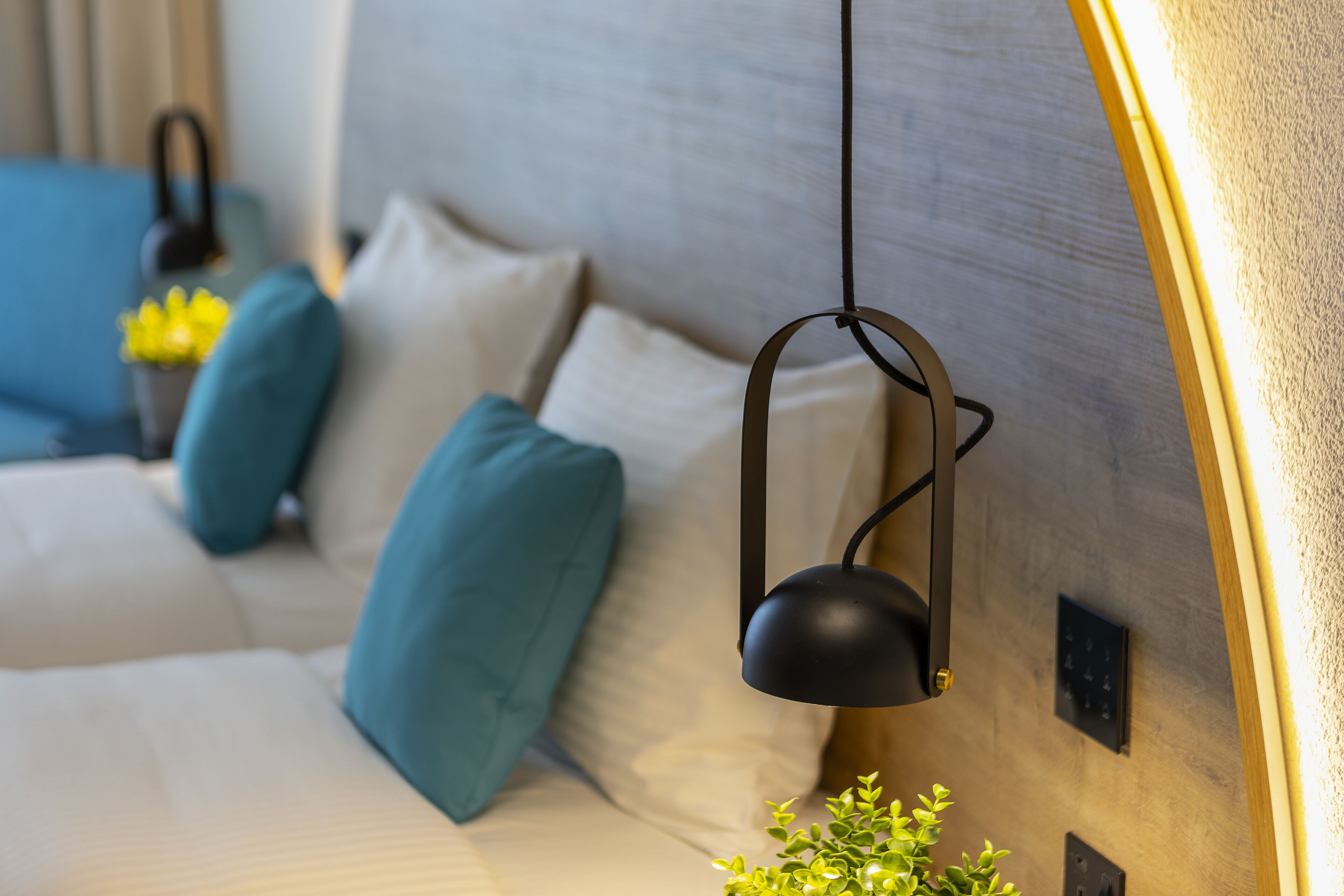 Quarto moderno com cama, almofadas verde-água, uma pequena planta e um abajur suspenso preto.