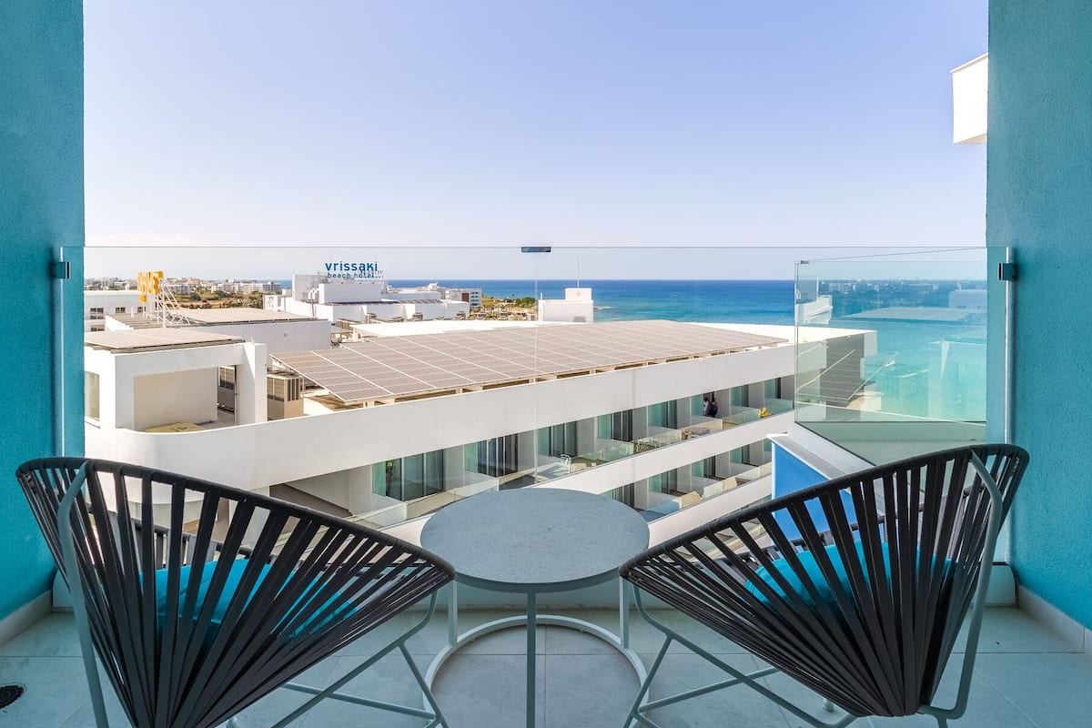 A balcony with two black wicker chairs and a small round table, overlooking a modern building with solar panels and a view of the sea.