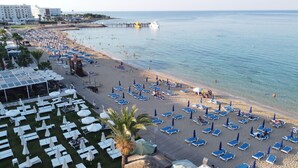 A beach with rows of sun loungers and umbrellas, a pier with boats, and a hotel with outdoor seating.