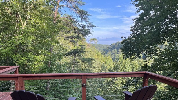 Una terraza de madera con dos sillas Adirondack y una mesa, con vistas a un valle boscoso.