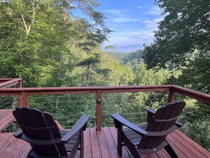 A wooden deck with two Adirondack chairs and a table, overlooking a forested valley.