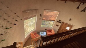 Une pièce aux hauts plafonds, avec un escalier en bois, une table ronde entourée de chaises et une vue sur l’extérieur grâce à de grandes fenêtres.