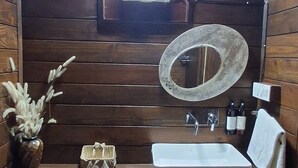 Une salle de bain aux murs lambrissés, avec un lavabo blanc, un miroir rond, un ventilateur et un petit panier décoratif.