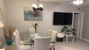 Uma sala de jantar com mesa de vidro, cadeiras brancas, um lustre, um quadro, uma televisão e uma porta.