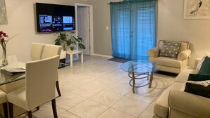 Uma sala de estar com televisão, ventilador de teto, mesa de centro de vidro e sofá.