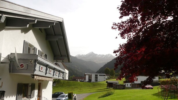 A residential building with a sloped roof, a balcony with outdoor furniture, and a view of mountains in the background.