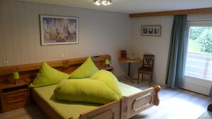 A bedroom with a wooden bed, green pillows, a bedside table with a lamp, a small round table with a chair, and a window with a view of greenery.