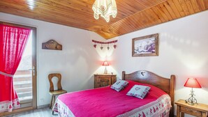 A bedroom with a wooden ceiling, a bed with a red bedspread, two bedside lamps, a wooden nightstand, a chair, and a window with red curtains.